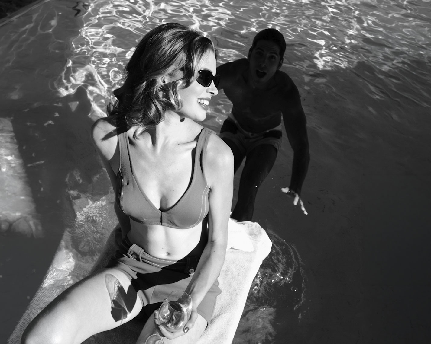 A woman holding a wine glass beside a swimming pool, with a man in the water